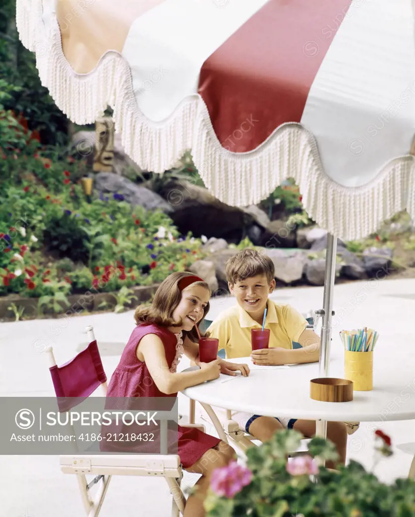 1970s BOY AND GIRL WITH COOL DRINKS AT TABLE WITH LARGE UMBRELLA OUTDOORS