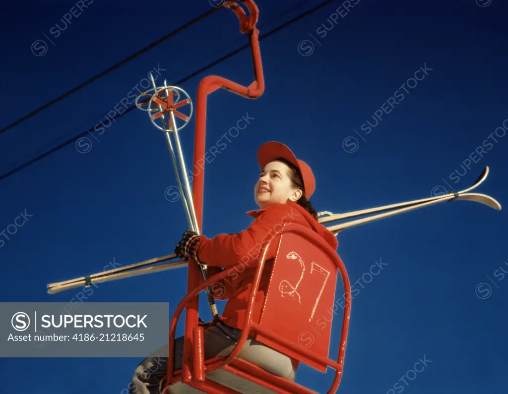 1950s BRUNETTE WOMAN RED SKI HAT JACKET SITTING IN RED SKI LIFT CHAIR HOLDING VINTAGE SKIS SKI POLES