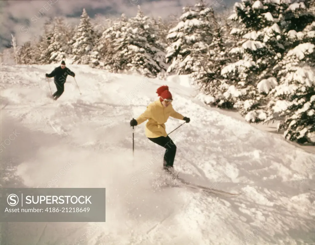 1960s 1970s TWO PEOPLE MAN WOMAN SKIING DOWN SNOWY SLOPE PINE TREES WOMAN WEARING YELLOW JACKET AND RED CAP