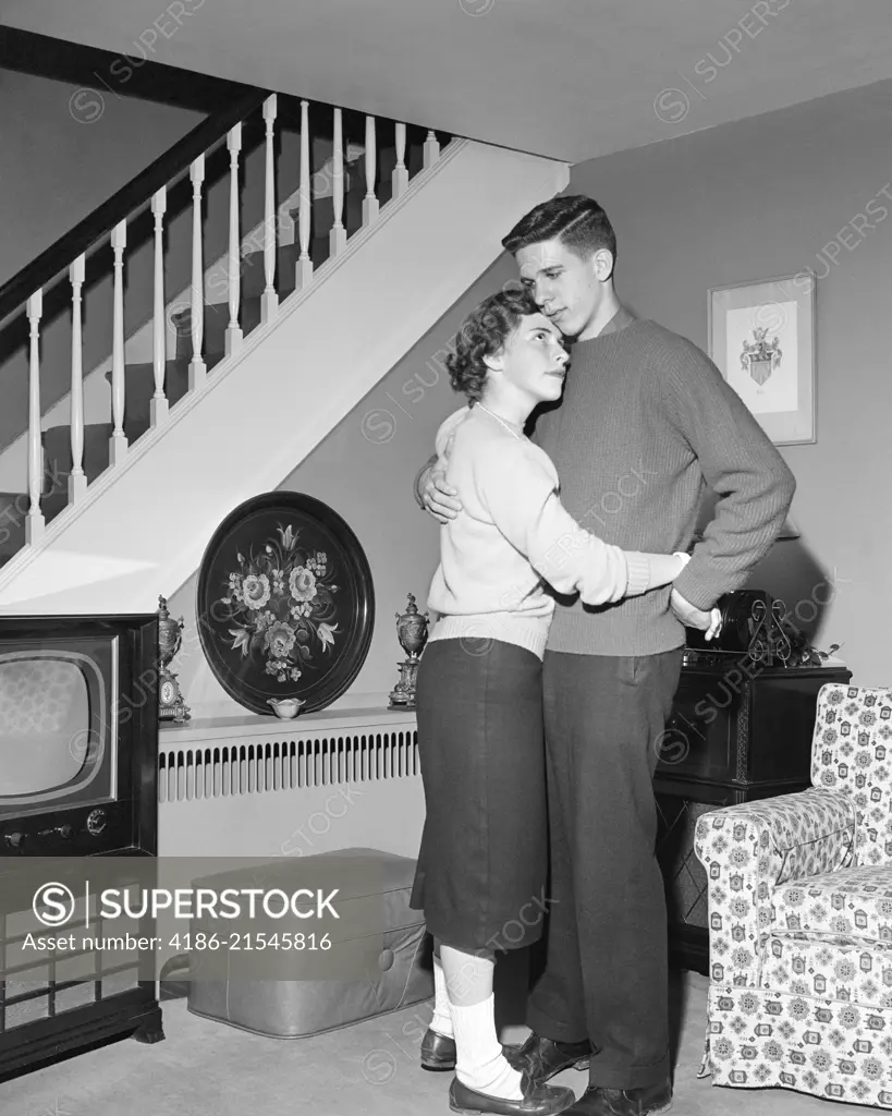 1950s HUGGING DANCING TEENAGE COUPLE IN LIVING ROOM