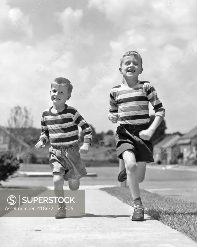 1950s TWO SMILING BOYS BROTHERS WEARING STRIPED SHIRTS SHORTS RUNNING ON SIDEWALK LOOKING AT CAMERA SUBURBAN NEIGHBORHOOD