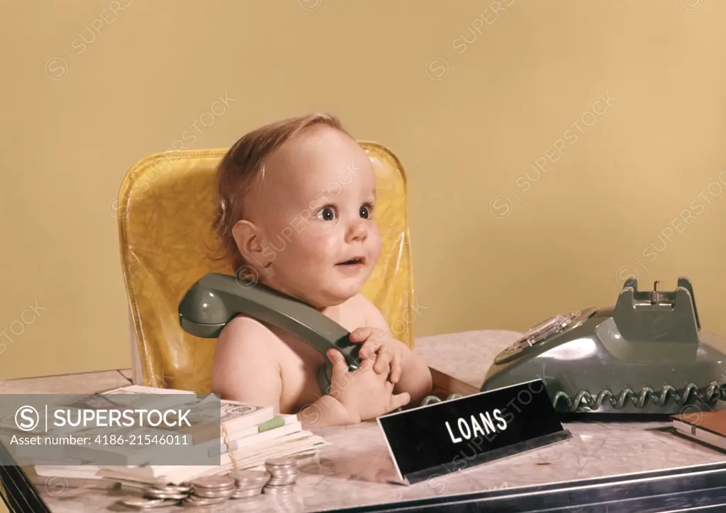 1960s WIDE EYED BABY SEATED AT LOAN OFFICER DESK HOLDING TELEPHONE