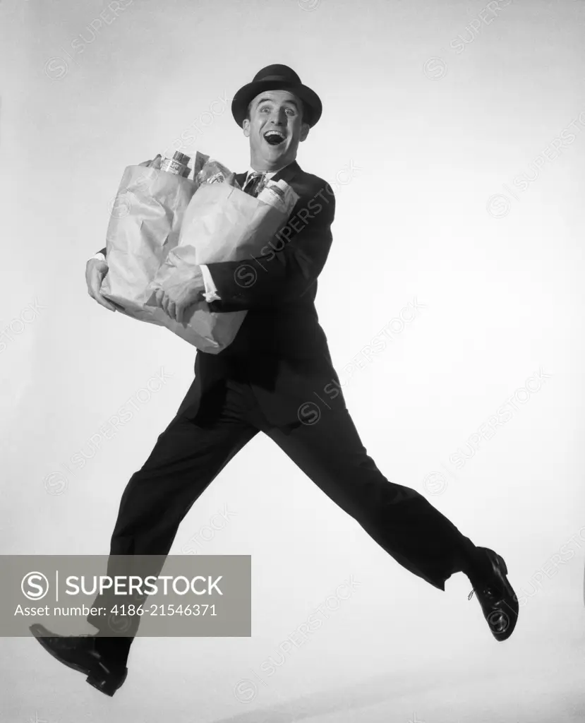 1950s 1960s HAPPY EXCITED MAN WEARING BUSINESS SUIT HAT CARRYING GROCERY SHOPPING BAGS RUNNING JUMPING IN AIR LOOKING AT CAMERA