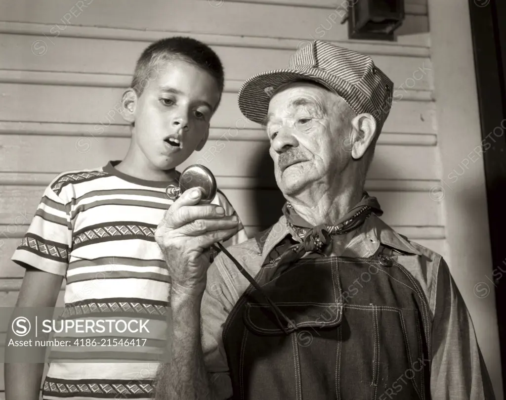 1950s ELDERLY MAN GRANDFATHER TRAIN ENGINEER HOLDING UP POCKET WATCH FOR BOY GRANDSON TO SEE