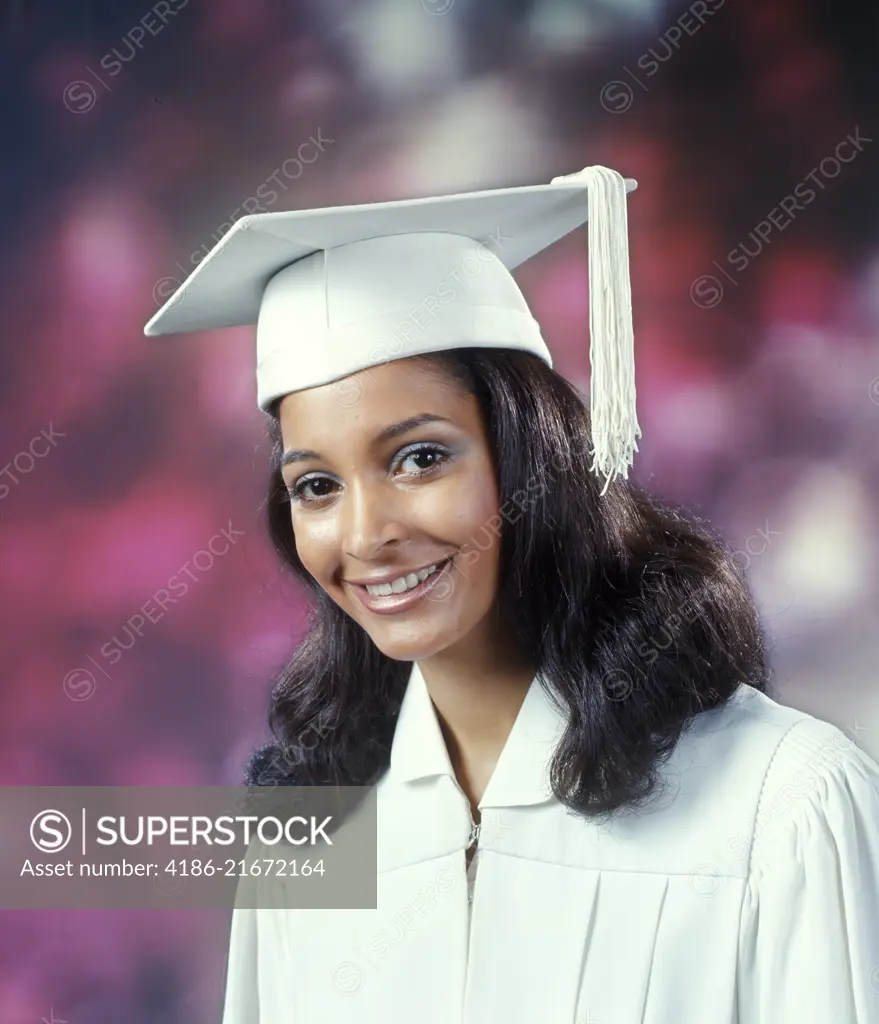 1970s YOUNG AFRICAN AMERICAN WOMAN GRADUATE IN WHITE CAP AND GOWN LOOKING AT CAMERA