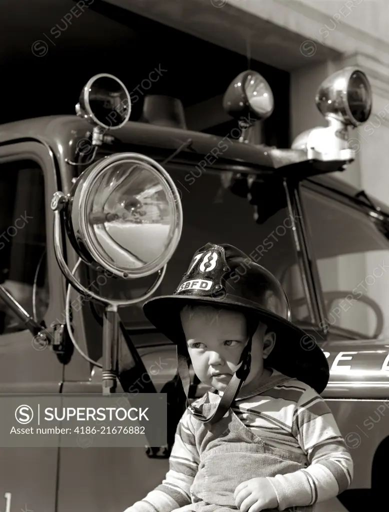 1950s 1960s BOY WEARING FIREMAN SAFETY HELMET IN FRONT OF FIRE TRUCK