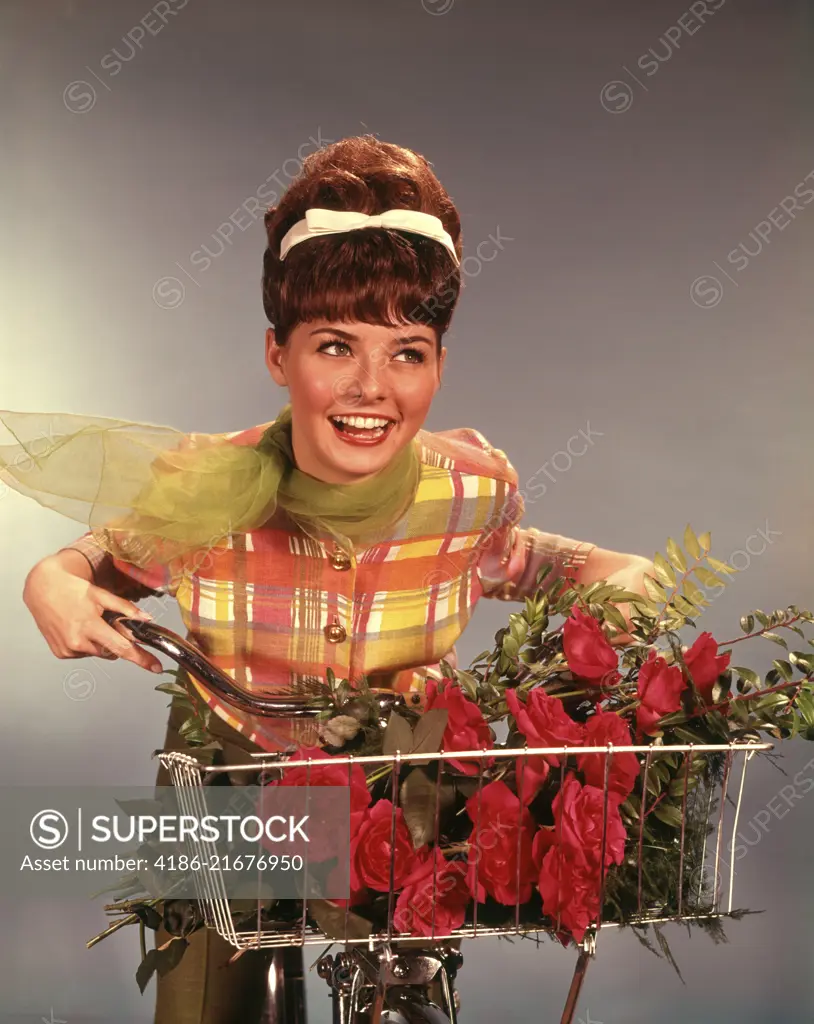 1960s SMILING TEEN GIRL WITH TEASED BRUNETTE BOUFFANT HAIRDO RIDING BICYCLE WITH RED ROSES BOUQUET IN BASKET