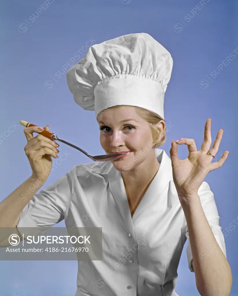 1960S WOMAN CHEF WEARING TOQUE LOOKING AT CAMERA TASTING FOOD MAKING OKAY SIGN SMILING