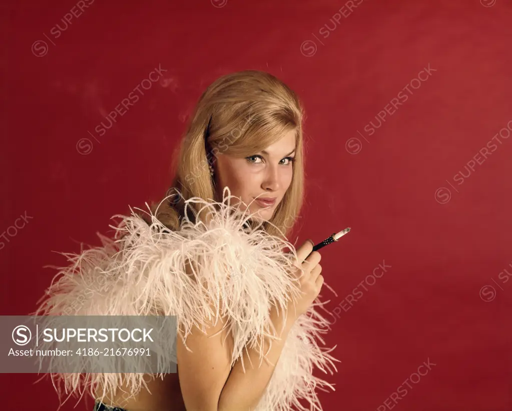 1960s SEXY BLONDE WOMAN LOOKING AT CAMERA SMOKING CIGARETTE IN LONG HOLDER WEARING WHITE FEATHER BOA AGAINST RED BACKDROP