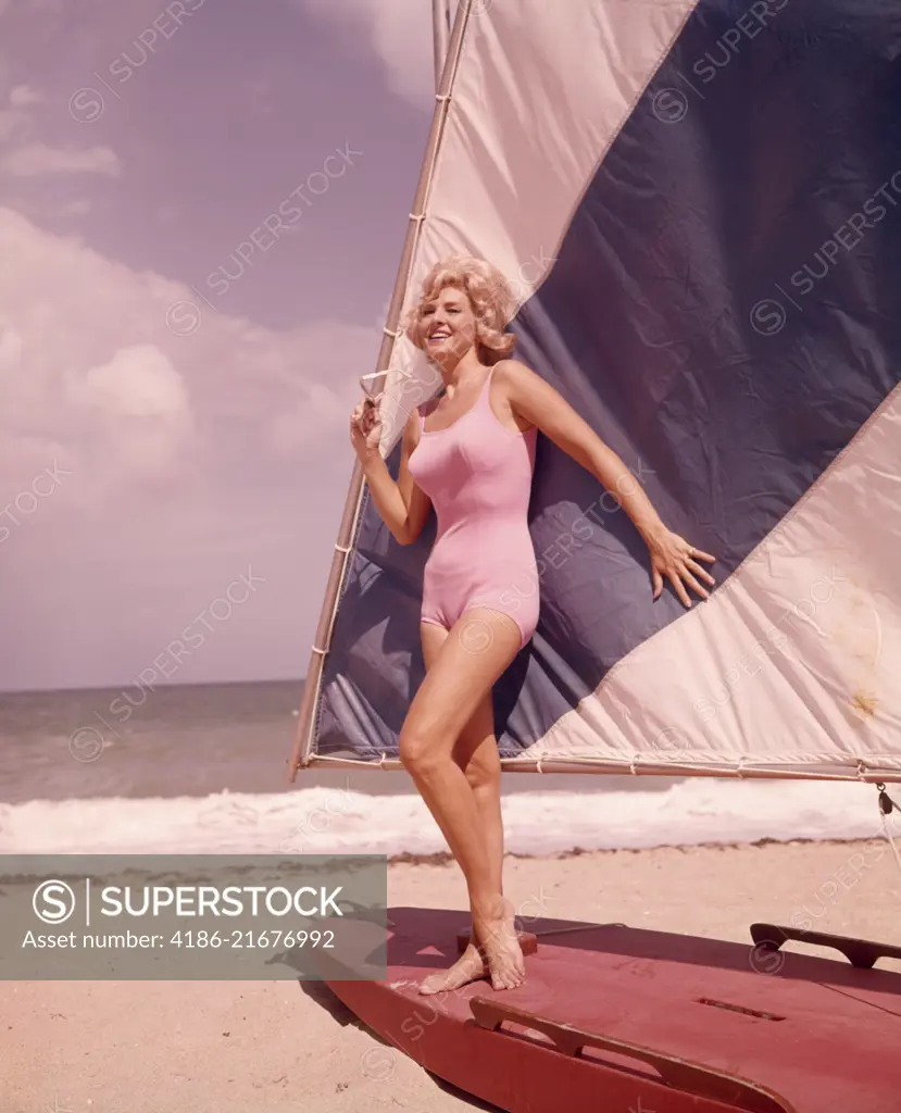 1960s BLOND WOMAN BEAUTY IN PINK SWIM WEAR BATHING SUIT POSED ON BLUE WHITE SAIL BOAT SAILFISH BOAT ON SUMMER SEASHORE BEACH