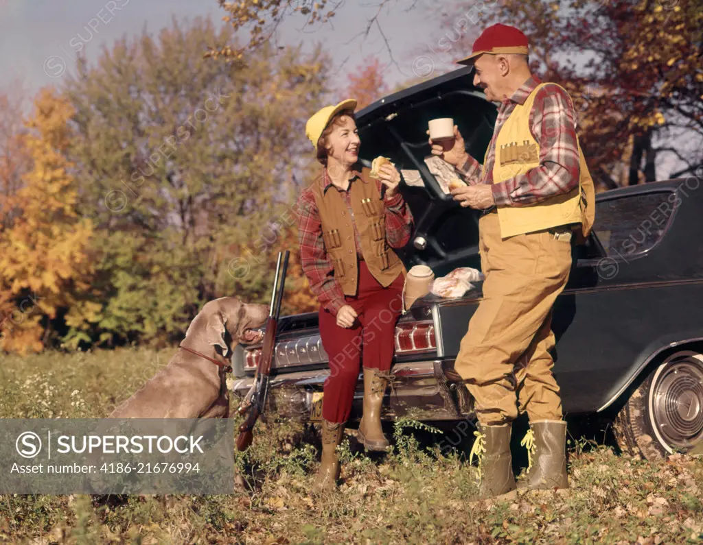 1970s ATTENTIVE WEIMARANER DOG WATCHING HUNTING COUPLE MAN AND WOMAN SITTING ON STANDING BY CAR TRUNK TALKING AND EATING LUNCH