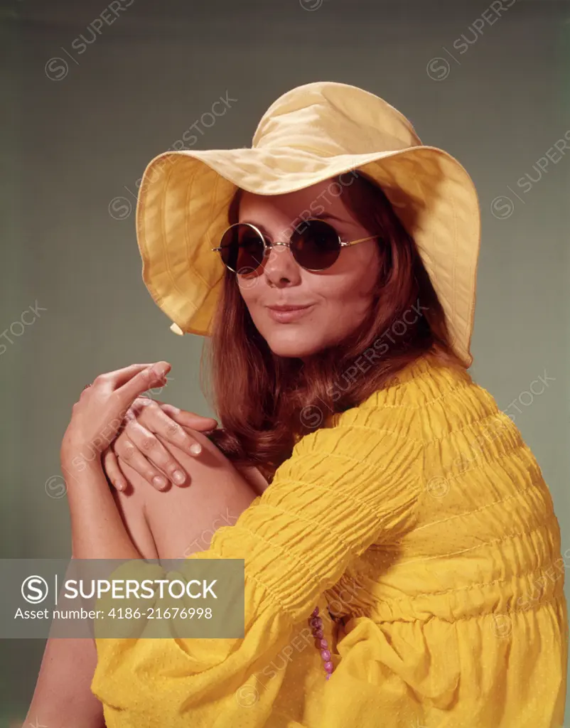 1960s PORTRAIT SMILING BRUNETTE TEENAGE WOMAN IN YELLOW FLOPPY BRIM HAT SMOCKED SHIRT ROUND LENS SUNGLASSES LOOKING AT CAMERA