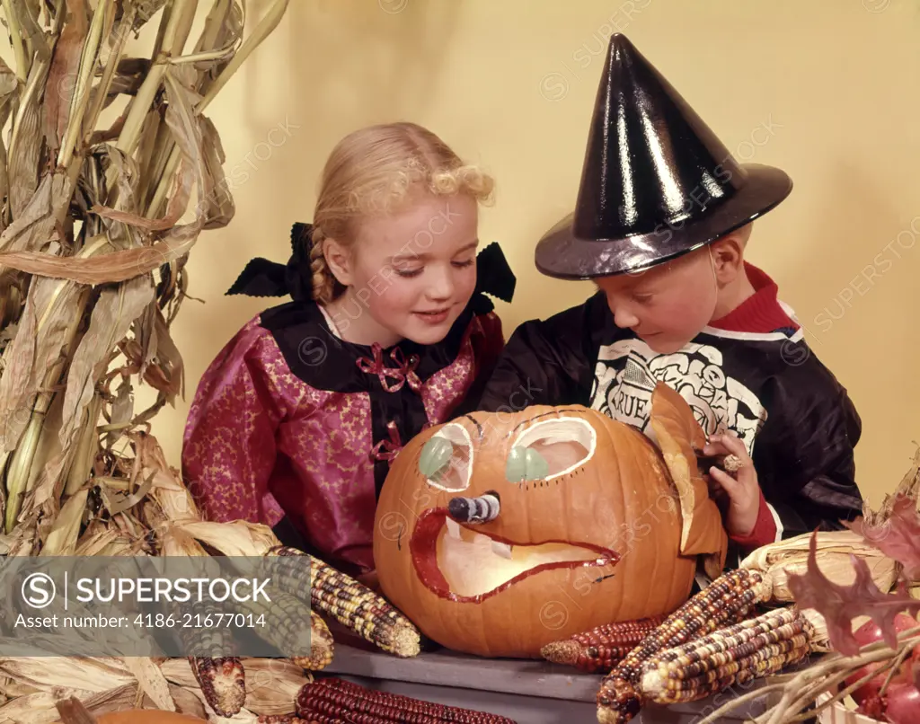 1960s TWO KIDS BLOND BROTHER AND SISTER WEARING HALLOWEEN COSTUMES LOOKING INTO CARVED JACK-O-LANTERN PUMPKIN