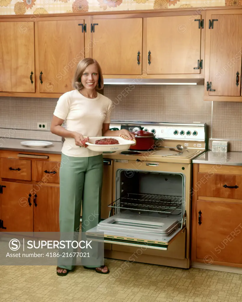 1970s SMILING BLOND WOMAN HOUSEWIFE LOOKING AT CAMERA HOLDING BAKED HAM STANDING BY STOVE OPEN OVEN DOOR