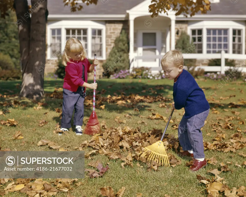 1960s BOY GIRL RAKING AUTUMN LEAVES IN YARD SUBURBAN HOUSE IN BACKGROUND 