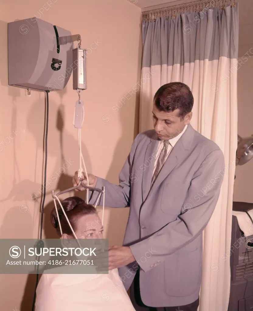 1950s 1960s MAN TECHNICIAN ADJUSTING TENSION ON WOMAN PATIENT CHIN BRACE NECK TRACTION MEDICAL EQUIPMENT