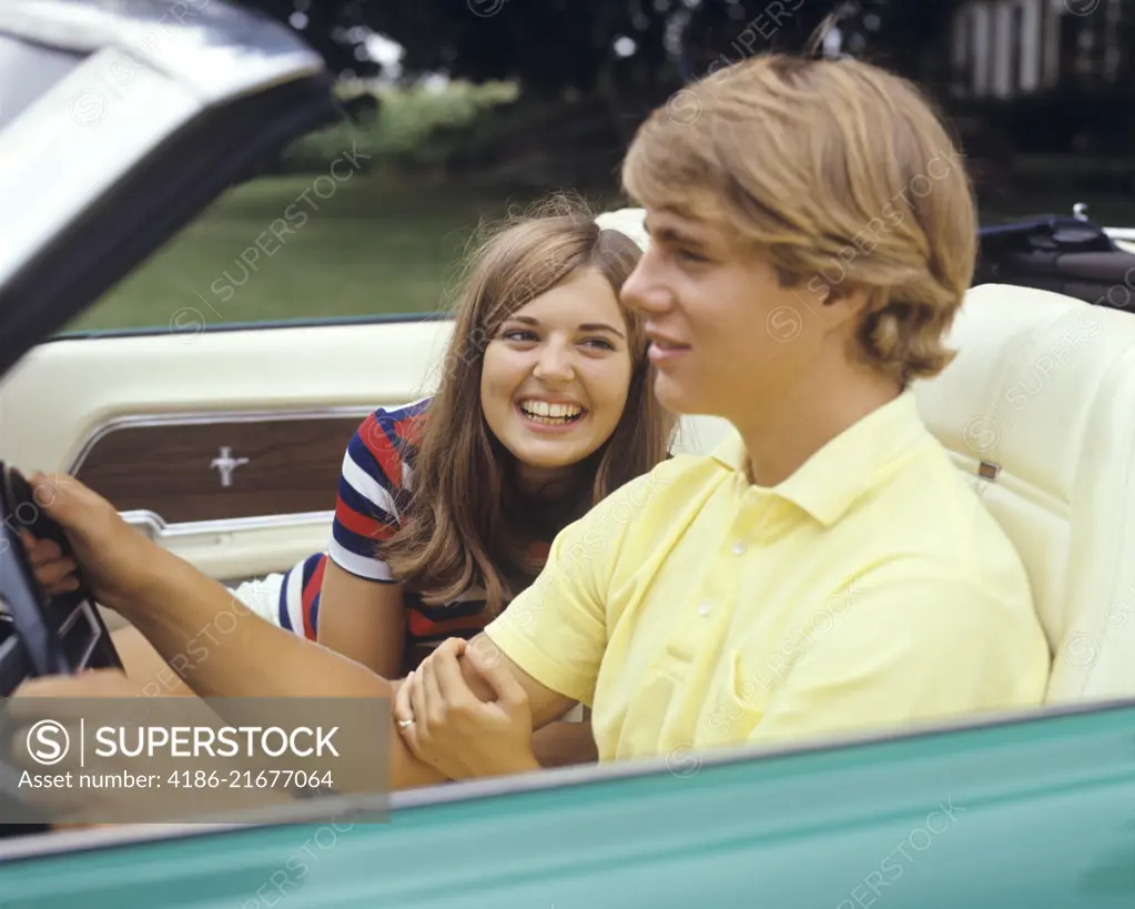 1960s 1970s SMILING TEENAGE COUPLE GIRL FLIRTING LOOKING AT HOLDING ARM OF BOY DRIVING CONVERTIBLE CAR 