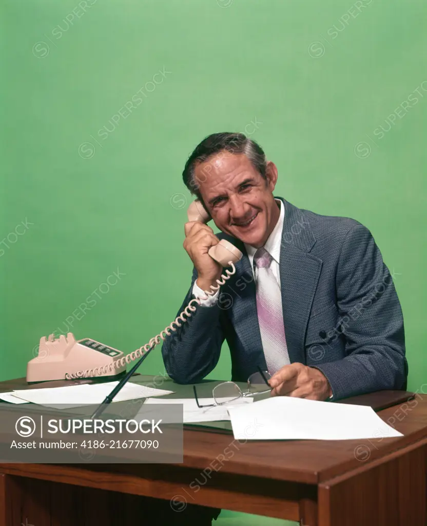 1970s SMILING MATURE MAN WEARING SUIT HOLDING GLASSES SITTING AT DESK SPEAKING ON TELEPHONE LOOKING AT CAMERA