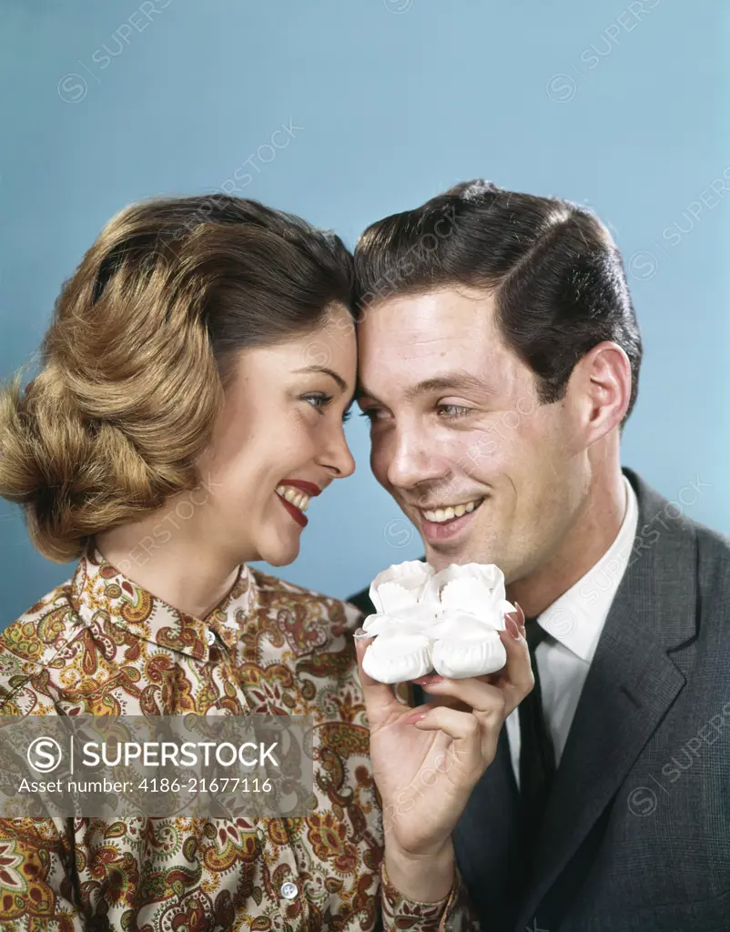 1960s SMILING COUPLE TOUCHING FOREHEADS LOOKING AT EACH OTHER HOLDING UP BABY SHOES 