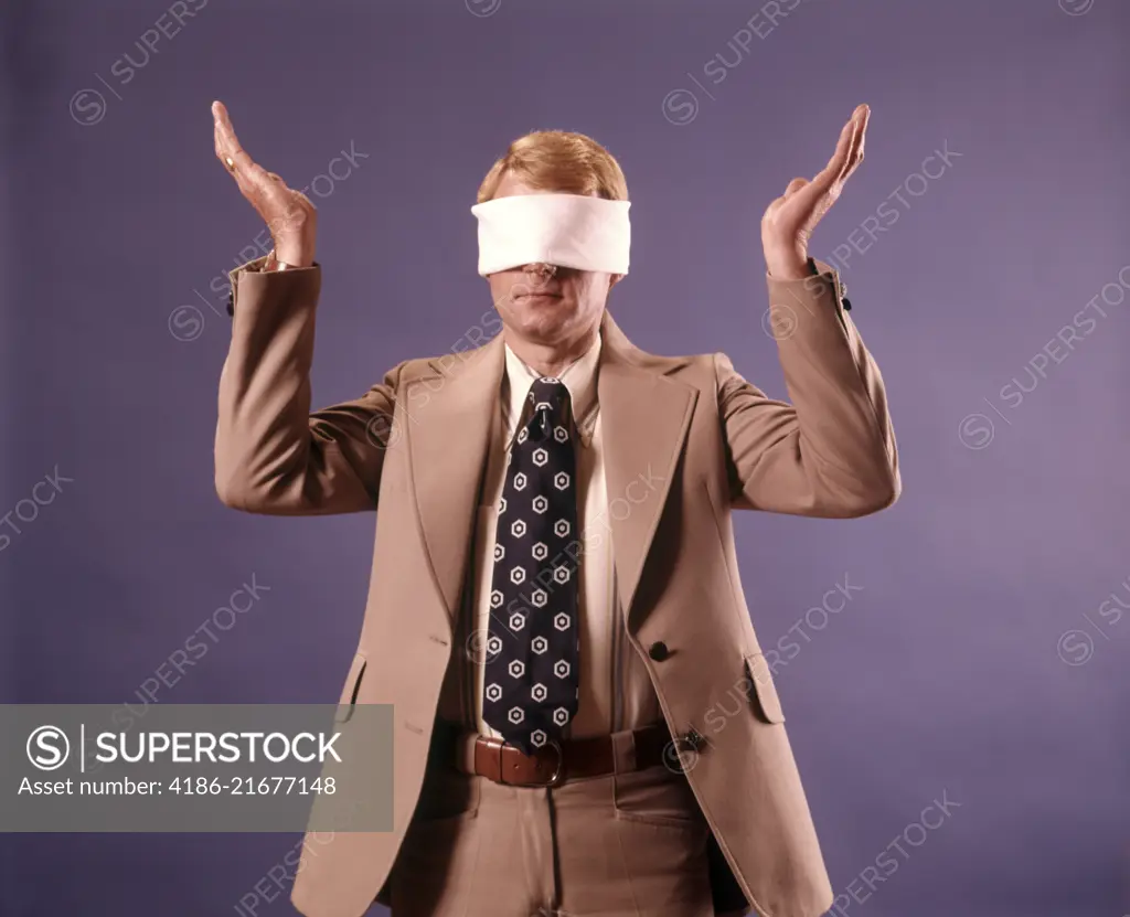 1970s EXECUTIVE MAN WEARING BUSINESS SUIT WEARING BLINDFOLD HOLDING BOTH HANDS UP IN HOPELESS GESTURE