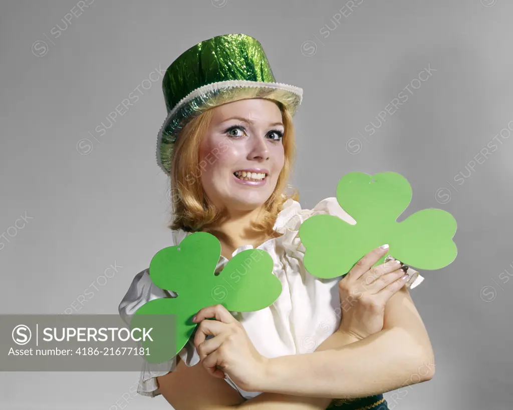 1960s SMILING BLOND WOMAN LOOKING AT CAMERA WEARING GREEN FOIL TOP HAT HOLDING TWO SAINT PATRICK DAY HOLIDAY IRISH SHAMROCKS 