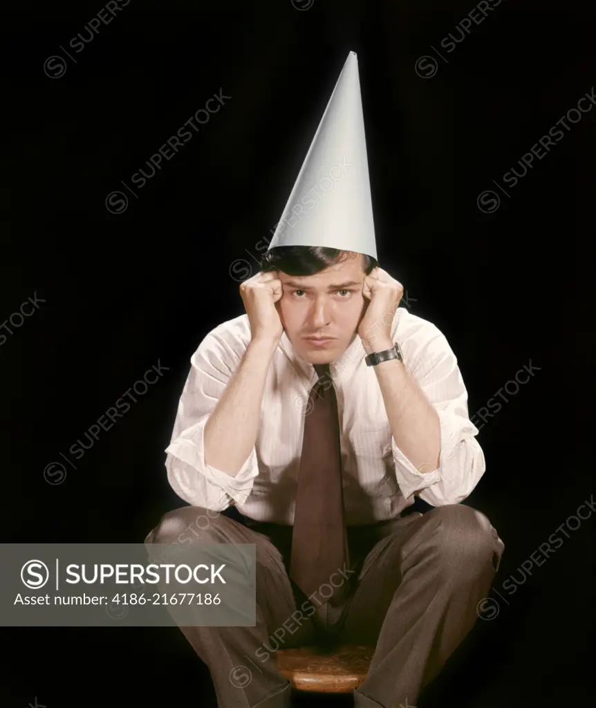 1960s UNHAPPY YOUNG MAN SITTING ON STOOL WEARING DUNCE CAP LOOKING AT CAMERA 