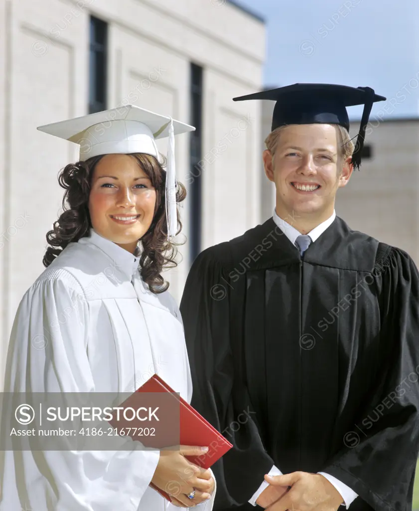 1970s PORTRAIT STUDENT COUPLE IN GRADUATION ROBES LOOKING AT CAMERA 