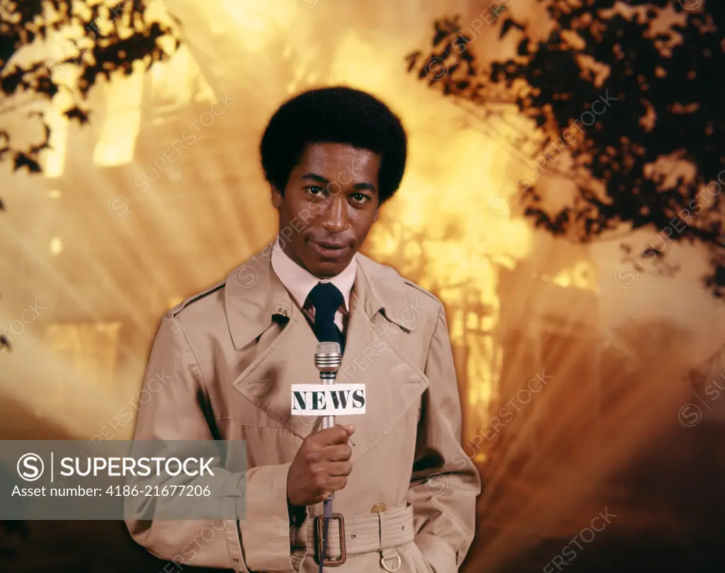 1970s AFRICAN AMERICAN MAN TELEVISION NEWS REPORTER WEARING RAINCOAT SPEAKING INTO MICROPHONE AT FIRE SCENE LOOKING AT CAMERA