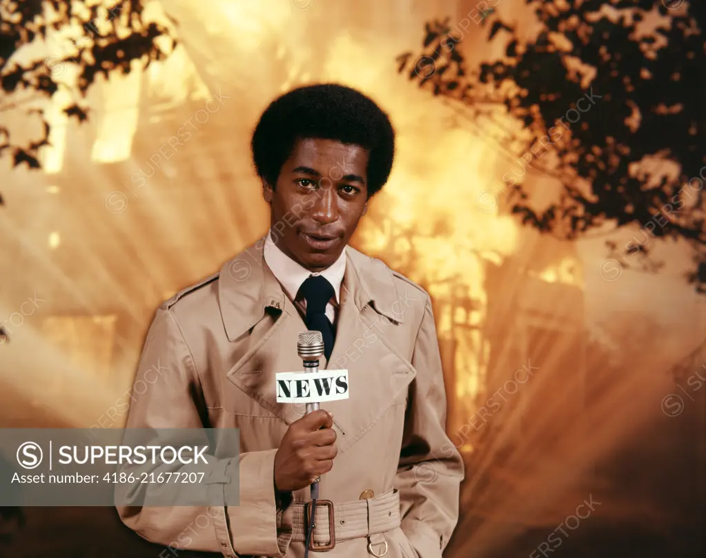 1970s AFRICAN AMERICAN TV NEWSMAN WEARING TRENCH COAT LOOKING AT CAMERA HOLD MICROPHONE REPORTING AT SCENE OF RAGING HOUSE FIRE