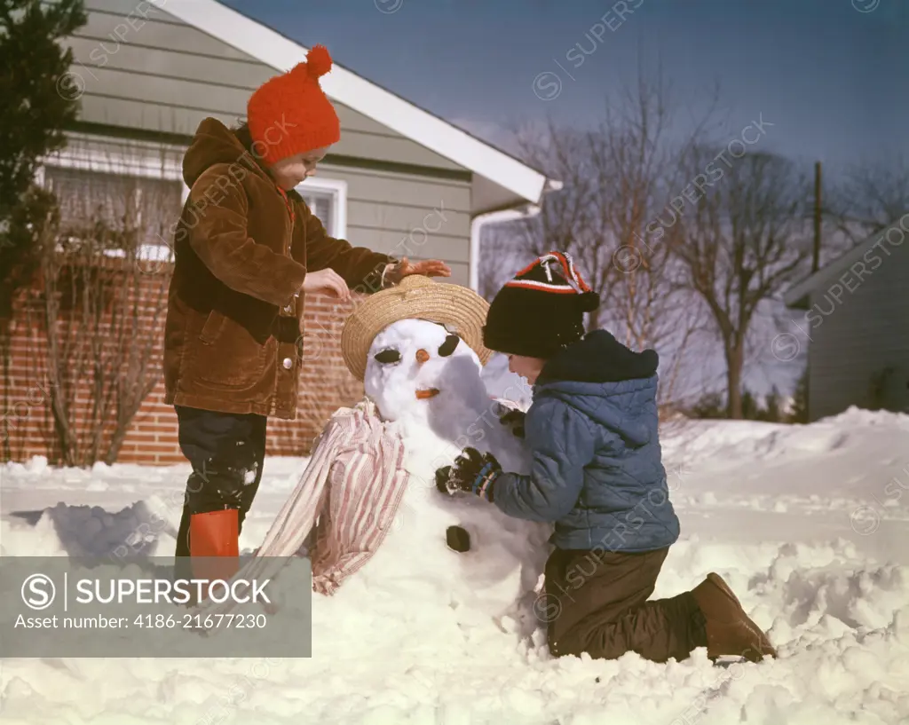 1960s 1970s BOY AND GIRL BROTHER AND SISTER MAKING SNOW MAN IN SUBURBAN HOUSE FRONT YARD