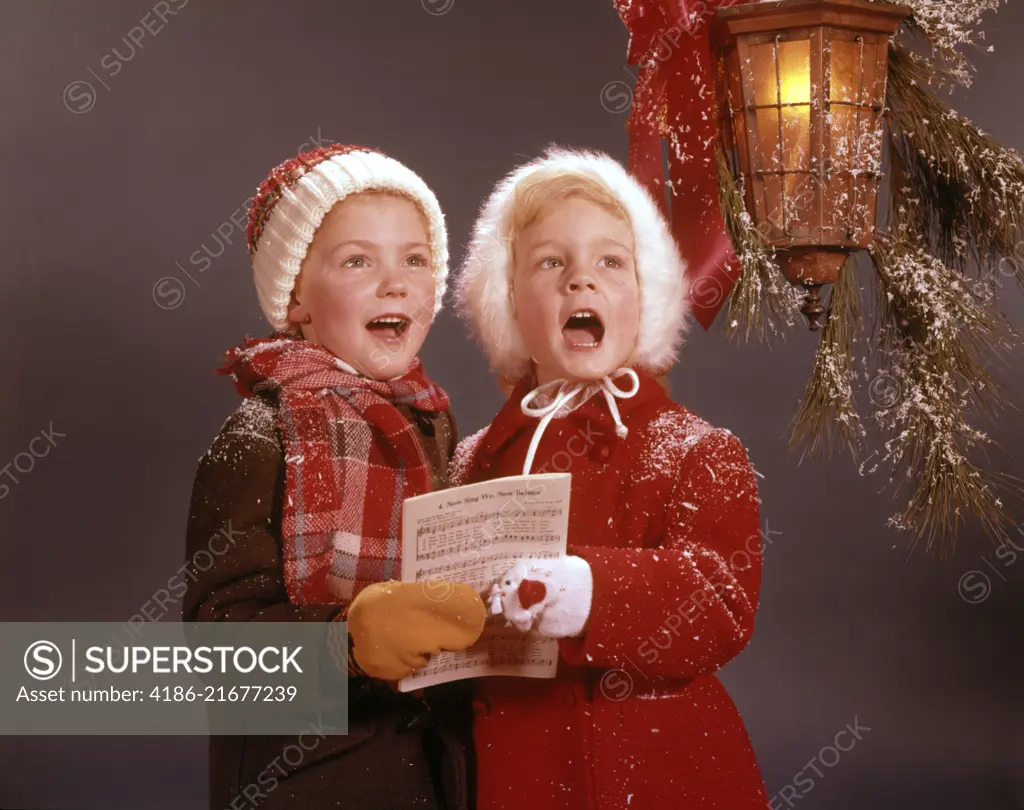 1960s TWO YOUNG CHILDREN BOY AND GIRL WINTER COATS SINGING CAROLS UNDER CHRISTMAS DECORATED CANDLE LANTERN