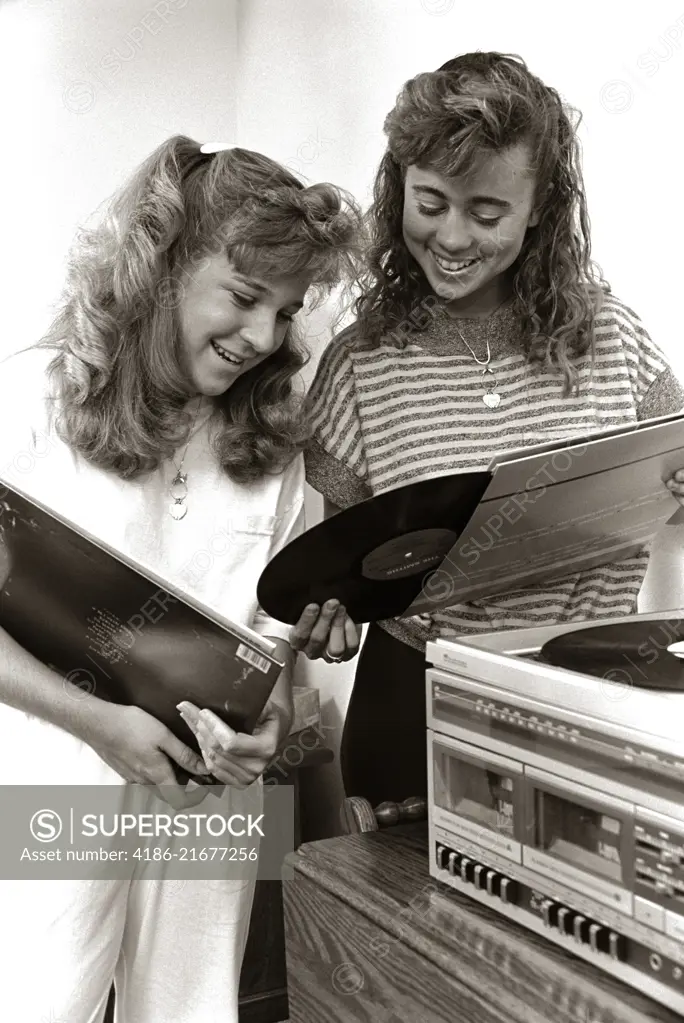 1980s 2 TEENAGE GIRLS READING VINYL RECORD ALBUM JACKET COVERS LISTENING TO LONG PLAYING PHONOGRAPH PLAYER