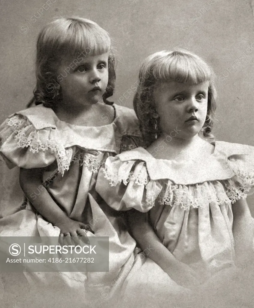 1890s PORTRAIT TURN OF THE 20TH CENTURY TWIN GIRLS SISTERS WEARING IDENTICAL DRESSES AND SHOWING MOROSE SAD FACIAL EXPRESSIONS