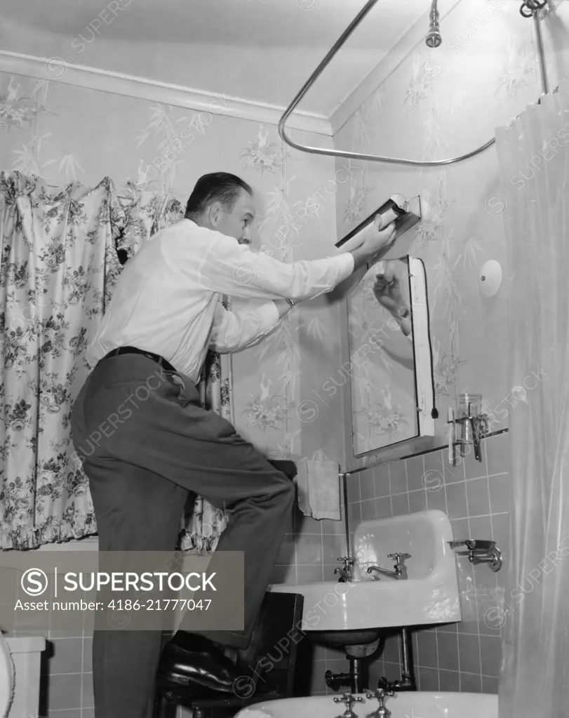 1950s MAN ON HOME REPAIR STEP STOOL IN BATHROOM REPLACING FLORESCENT LIGHTING FIXTURE ABOVE SINK