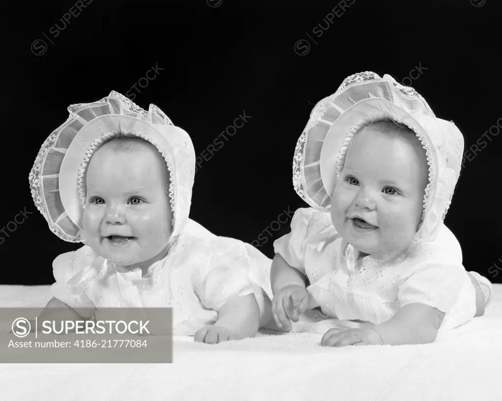 1950s TWIN BABY GIRLS WEARING BONNETS LYING ON THEIR STOMACHS SMILING