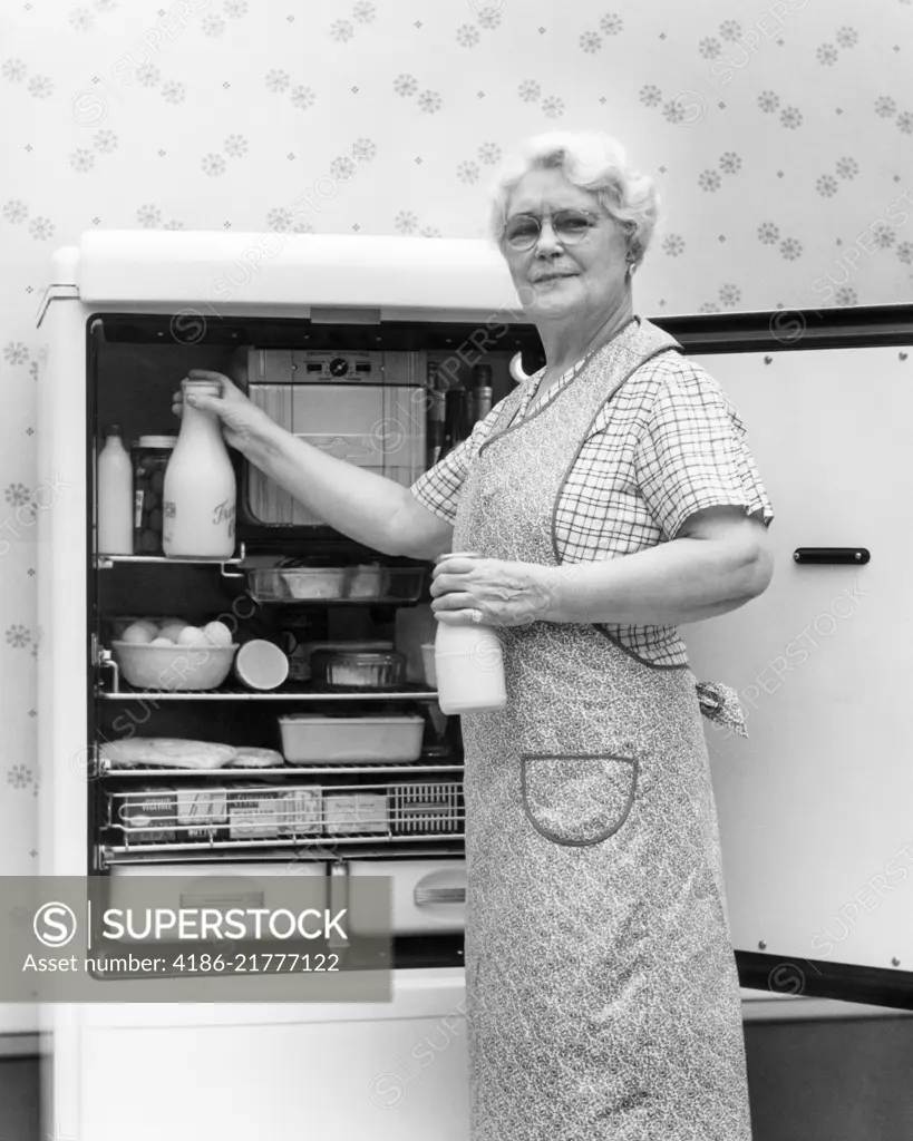 1930s 1940s SENIOR WOMAN HOUSEWIFE GRANDMOTHER LOOKING AT CAMERA TAKING TWO GLASS BOTTLES OF MILK OUT OF KITCHEN REFRIGERATOR