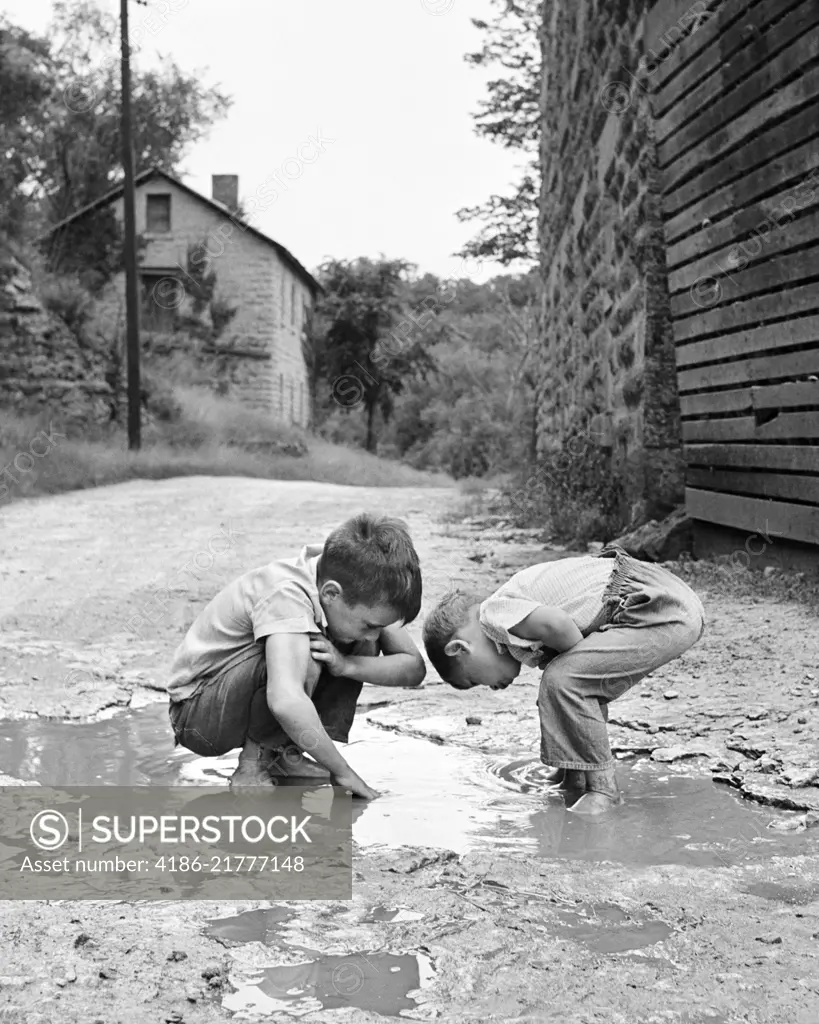 1950s 1960s TWO BOYS BENDING LOOKING AT PLAYING IN MUD PUDDLE