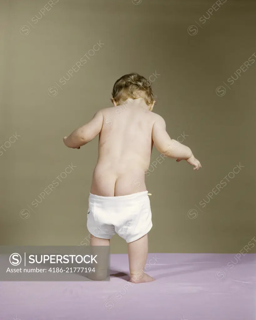 1960s REAR VIEW OF STANDING BABY WITH DIAPER FALLING DOWN SHOWING BARE BOTTOM