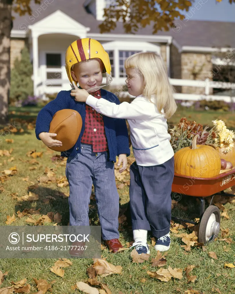 1960s BLONDE GIRL WEARING SADDLE OXFORDS HELPING BOY HOLDING FOOTBALL PUT ON HELMET AUTUMN YARD RED WAGON