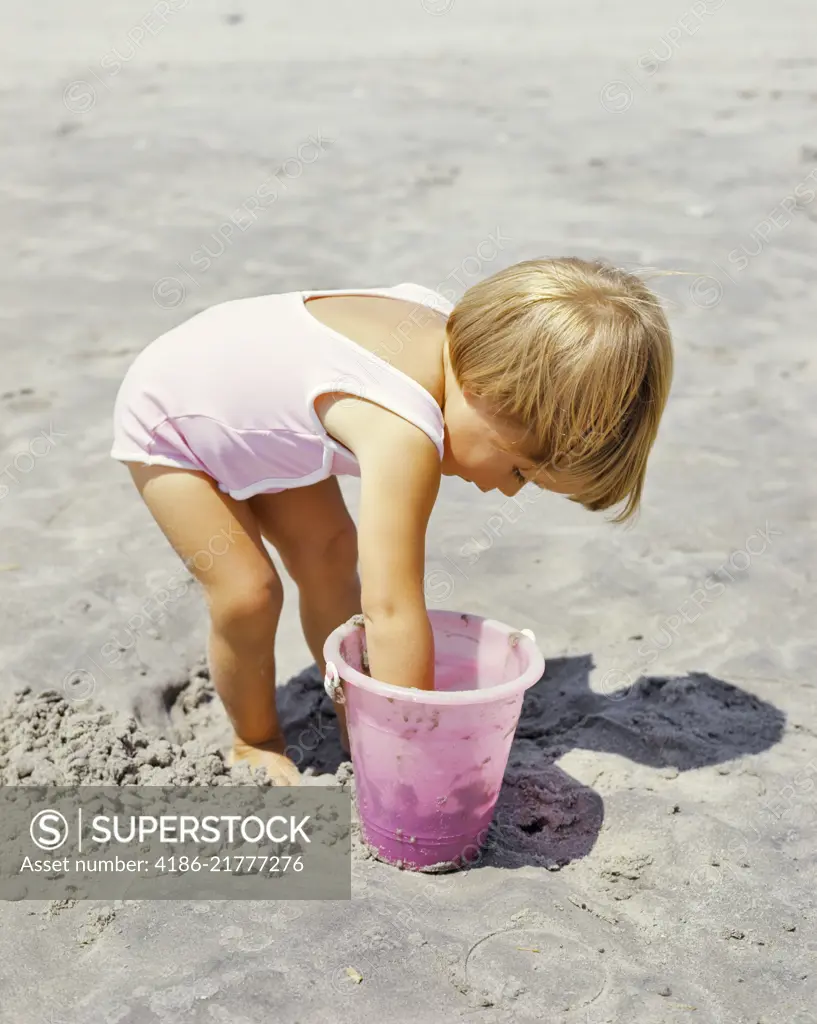 1970s LITTLE BLOND GIRL PINK BATHING SUIT PLAYING IN SAND WITH PINK PLASTIC PAIL ON BEACH