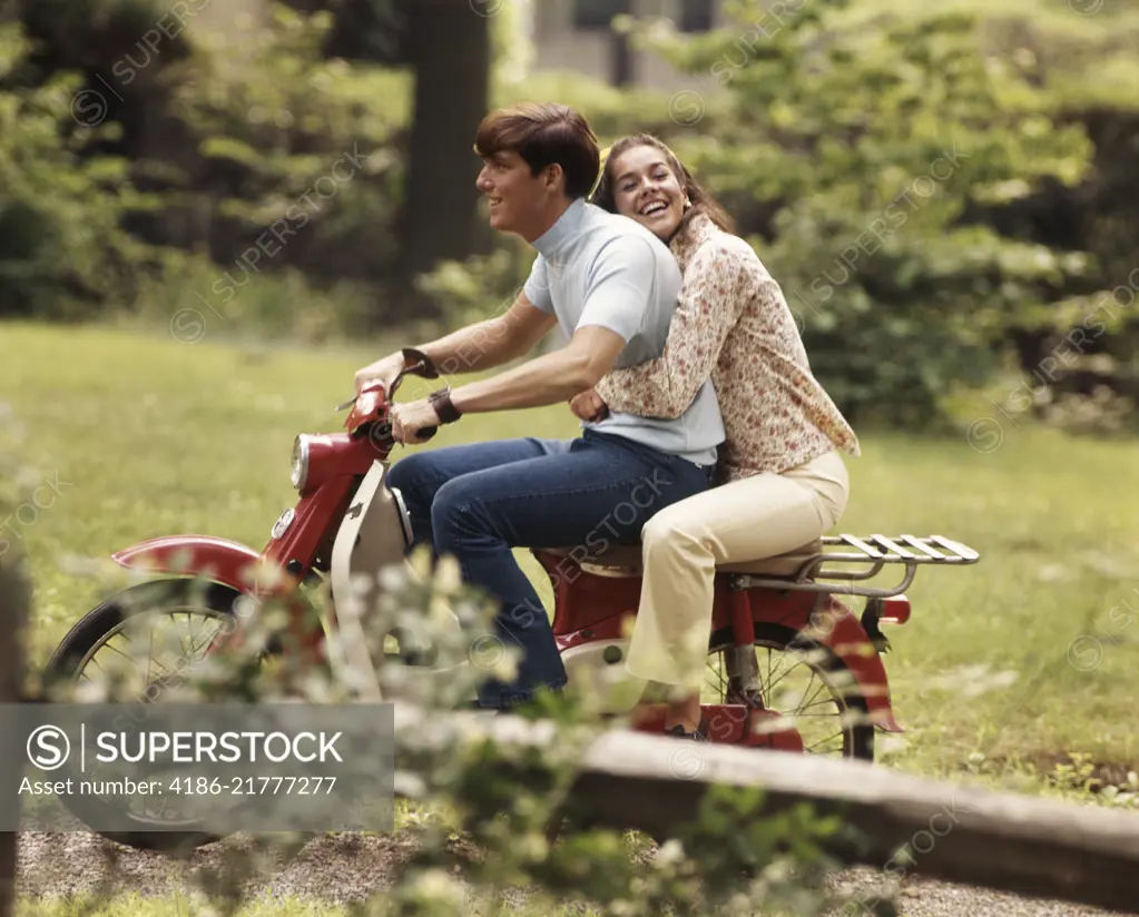 1960s 1970s YOUNG TEENAGE COUPLE RIDING ON MOTORBIKE BOY DRIVING GIRL HOLDING ON BEHIND LAUGHING