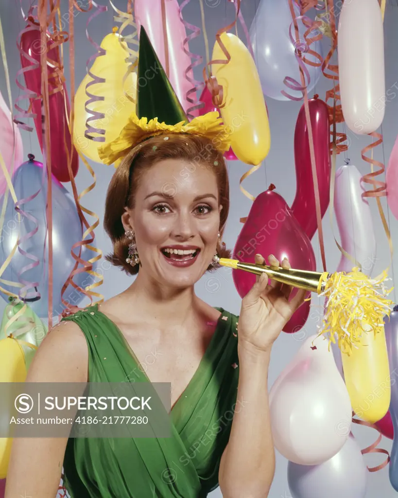 1960s SMILING WOMAN LOOKING AT CAMERA AMID STREAMERS AND BALLOONS HOLDING NEW YEARS NOISE MAKER