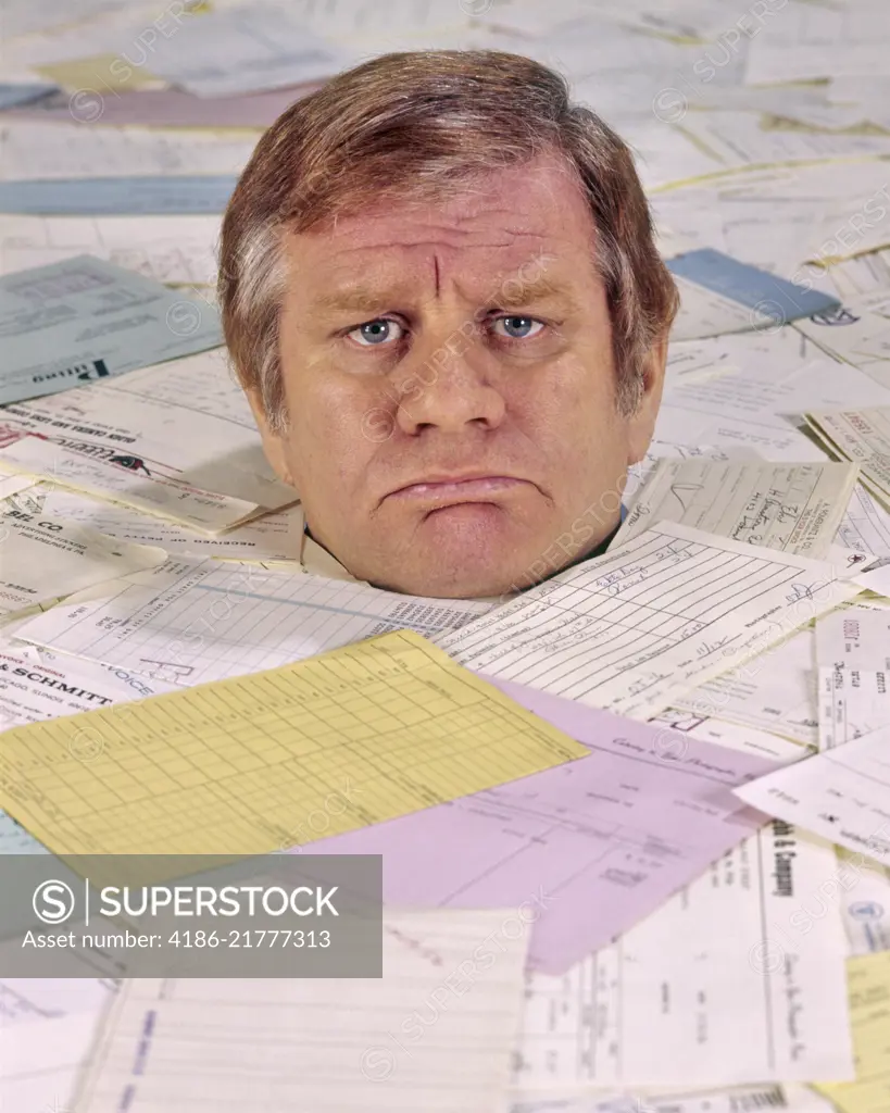 1970s 1980s ANGRY UNHAPPY FRUSTRATED MAN LOOKING AT CAMERA BURIED UP TO HIS NECK IN BILLS INVOICES PAPER WORK