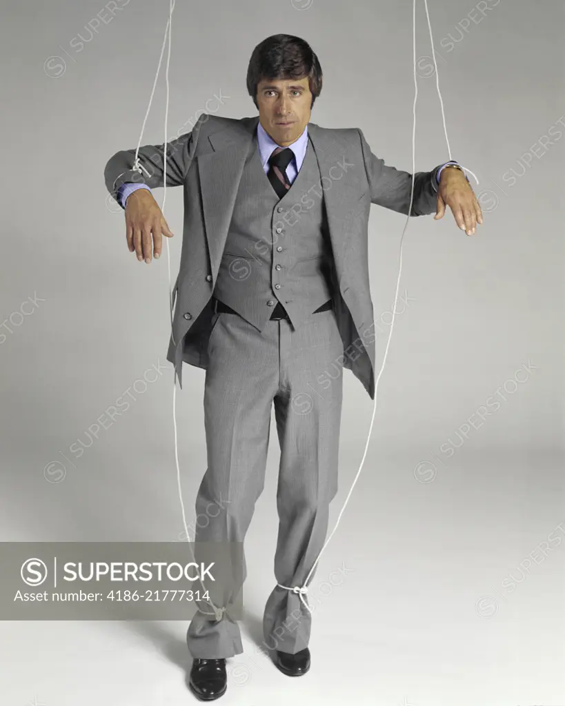 1970s BUSINESSMAN SUSPENDED ON PUPPET STRINGS LOOKING AT CAMERA