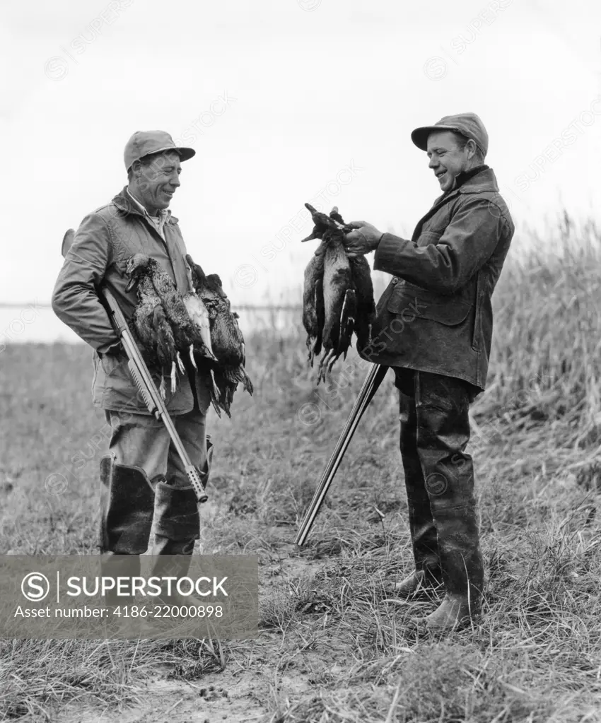 1950s TWO MEN DUCK HUNTERS WITH SHOTGUNS ADMIRING A BRACE OF DUCKS