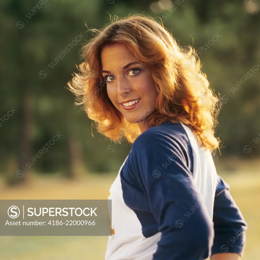 1970s 1980s SMILING YOUNG WOMAN SUNLIGHT ON HER GOLDEN HAIR LOOKING AT CAMERA OVER HER SHOULDER