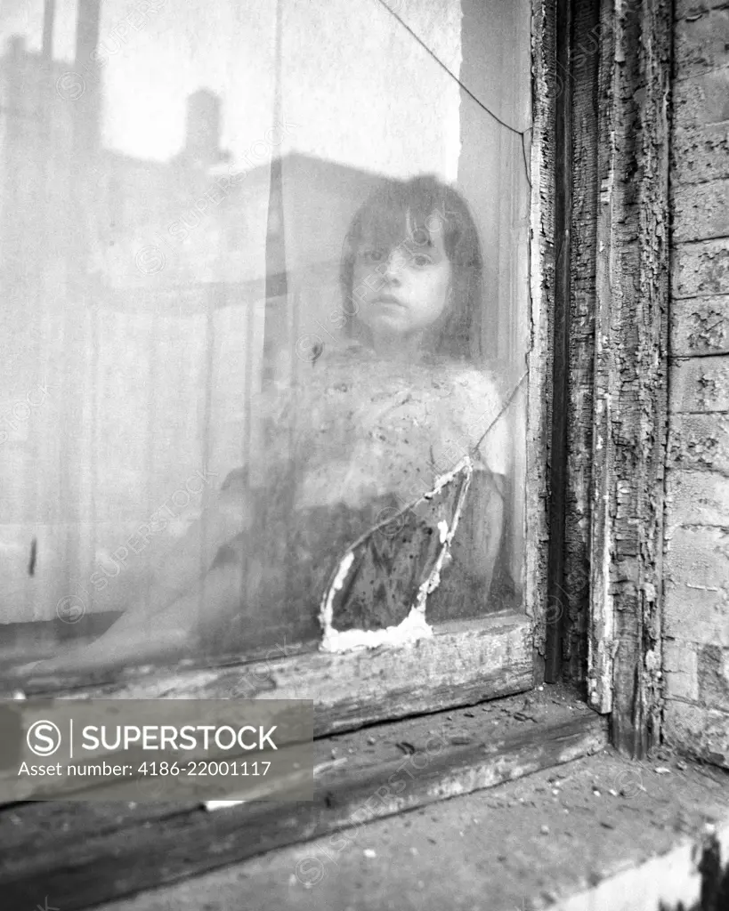 1970s POOR LITTLE GIRL LOOKING AT CAMERA OUT OF BROKEN WINDOW OF SLUM APARTMENT