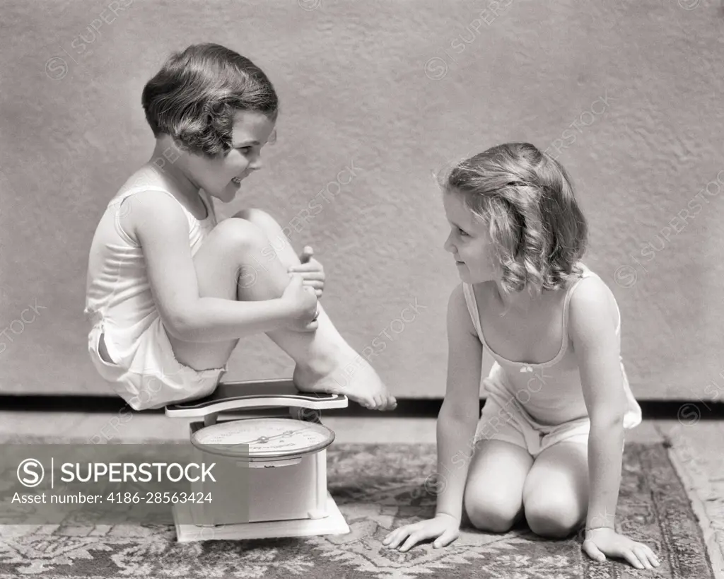 1930s TWO SMILING LITTLE GIRLS WEARING COTTON UNDERSHIRTS AND PANTIES  TALKING TOGETHER ONE SITTING ON AN ANTIQUE WEIGHT SCALE - SuperStock