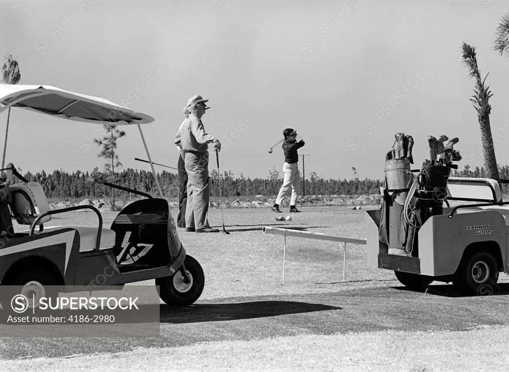 Three Men Playing Golf Teeing Off From Tee With Driver Near Golf Carts Outdoor 1960S