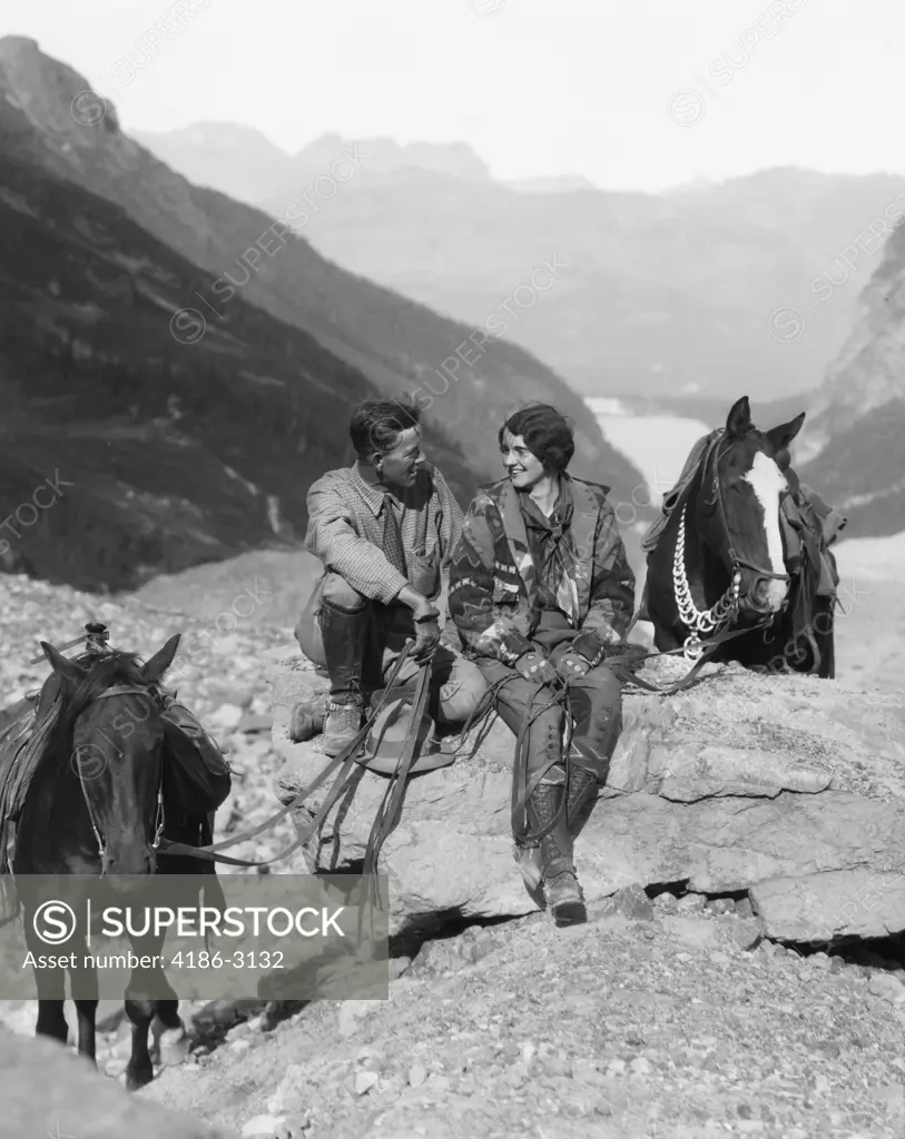 1920S 1930S Couple Man Woman Wearing Riding Gear Jodhpurs Boots Spurs Sitting On Large Rock By Two Horses With Western Saddles